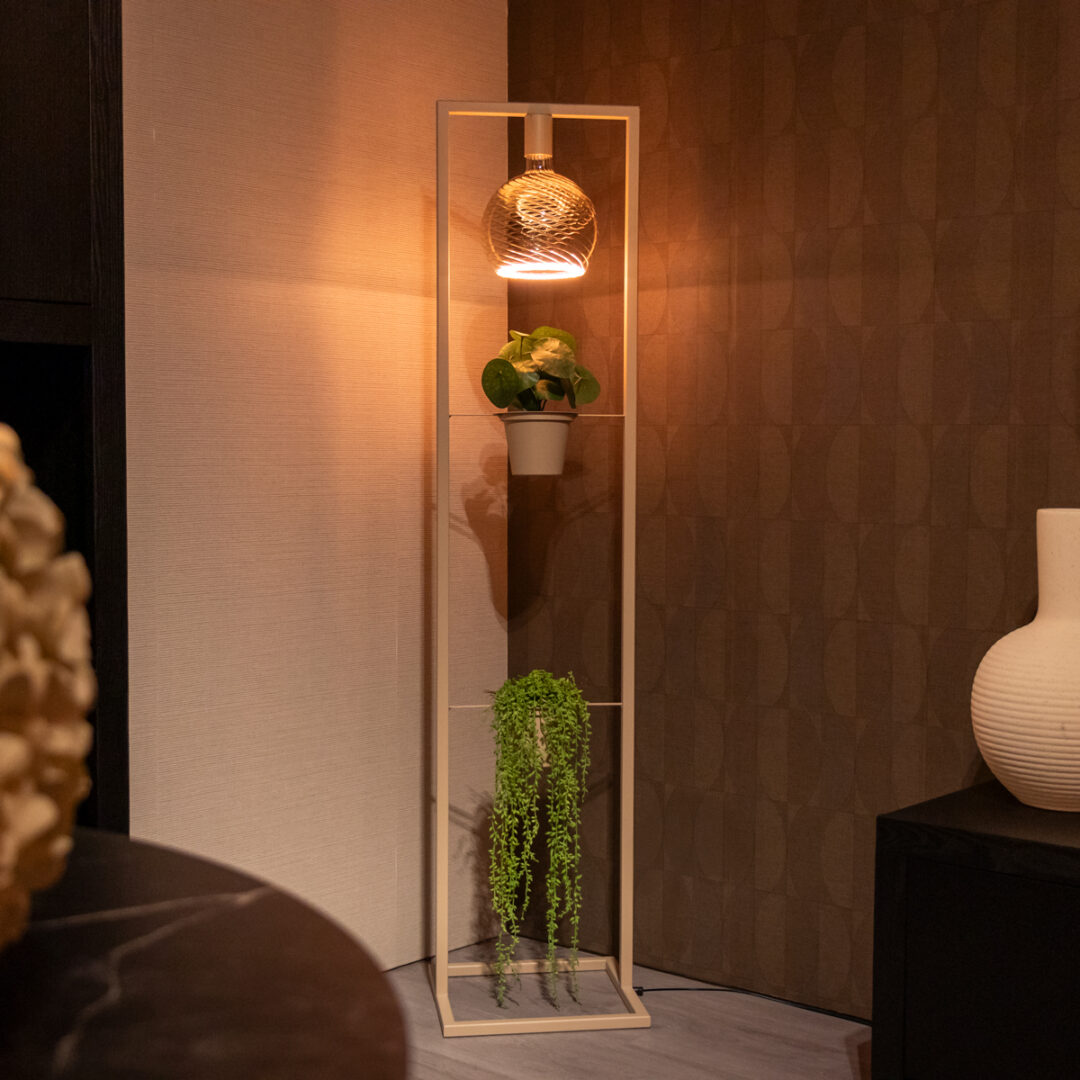 Beige staande lamp van Straluma met geïntegreerd rek en losse lichtbron, sfeervol verlicht in een hoek van de woonkamer.