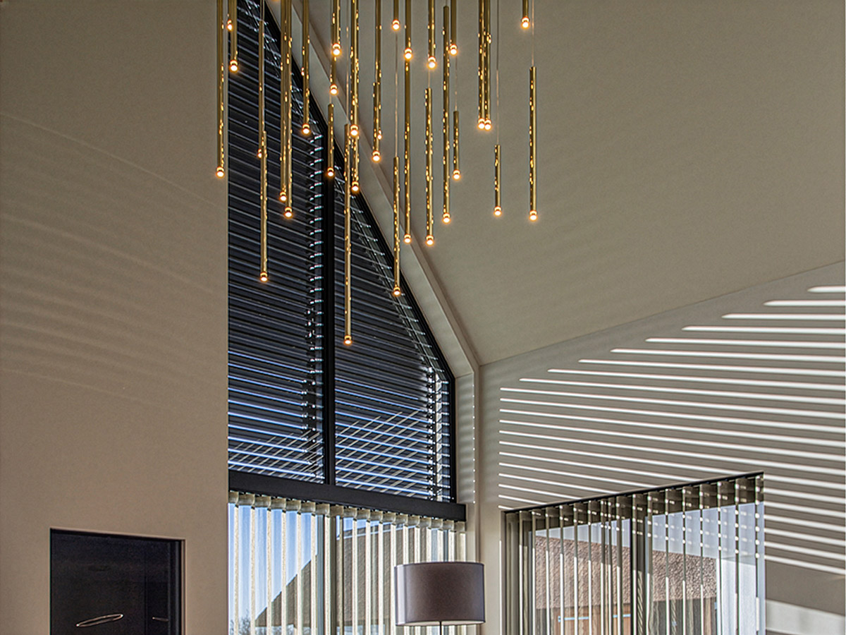 Moderne gouden hanglamp in hoge woonkamer met veel daglicht – Straluma verlichting.